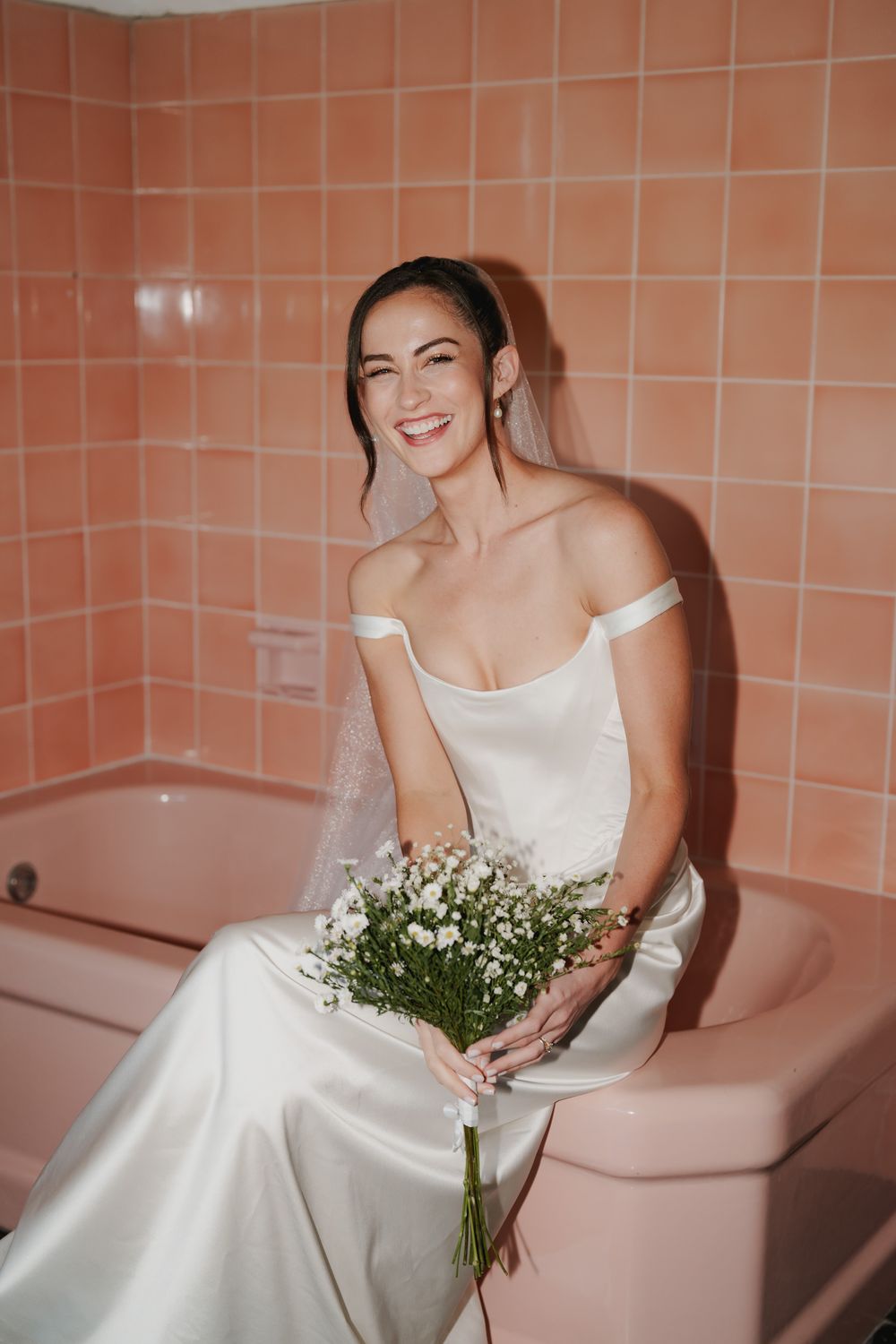 Smiling bride in an off-the-shoulder satin wedding gown and veil holds a white wildflower bouquet while sitting on a vintage pink bathtub.