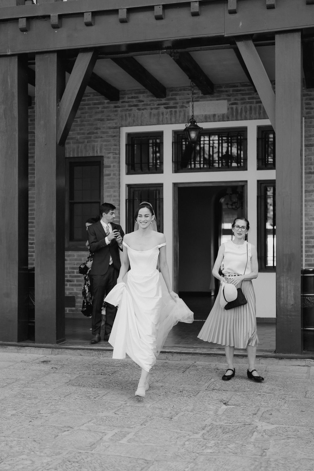 Black and white photo of a bride in an off-the-shoulder gown walking out of a brick building as a groom and a smiling guest look on.