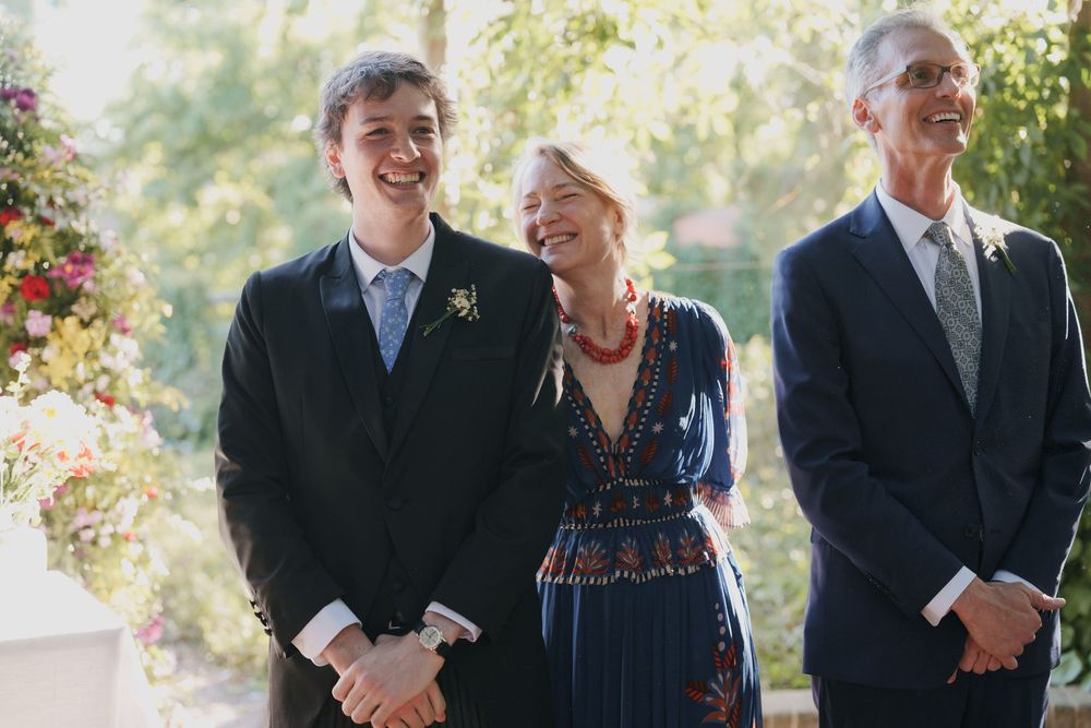 Smiling young man in a dark suit stands beside a woman in a floral dress and an older man in a navy suit at an outdoor wedding.