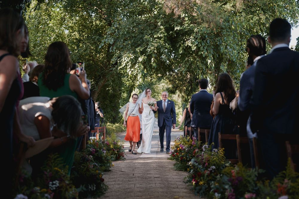 Bride in a white gown walks down a flower-lined brick aisle on her father's arm as seated wedding guests look on under leafy trees.