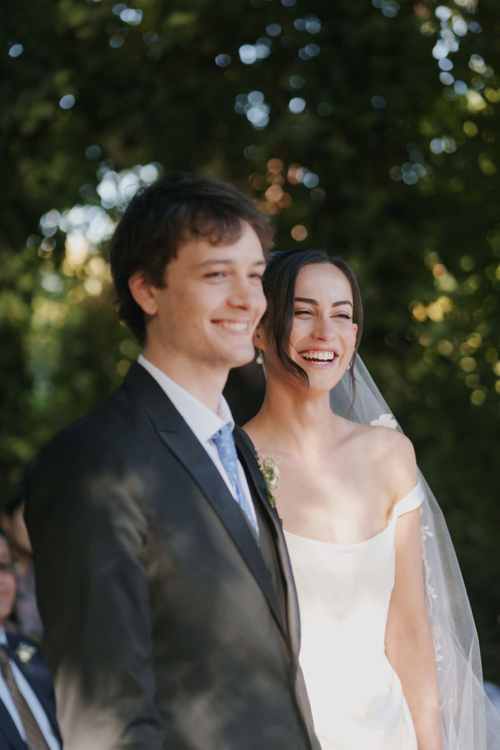 A smiling bride in a white gown and veil laughs beside a groom in a dark suit and blue tie at an outdoor wedding ceremony.
