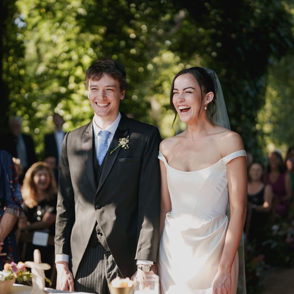 Laughing bride in an off-the-shoulder white gown and veil stands beside a smiling groom in a gray suit during an outdoor wedding ceremony.