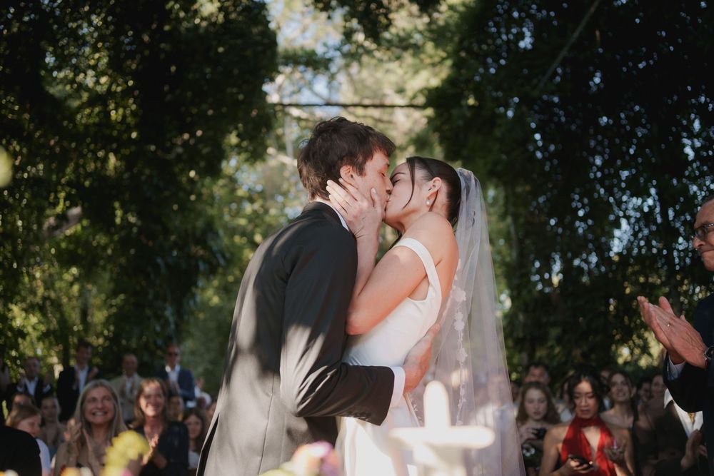 Bride in a white gown and veil kisses the groom in a gray suit during an outdoor wedding ceremony as guests watch and applaud under trees.
