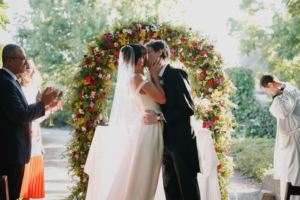 Bride and groom share a kiss under a floral wedding arch while guests applaud and an officiant looks on during an outdoor ceremony.