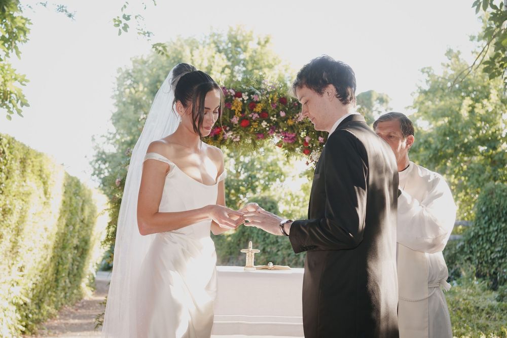 Bride in a white gown and veil exchanges rings with the groom under a floral arch during an outdoor garden wedding ceremony.