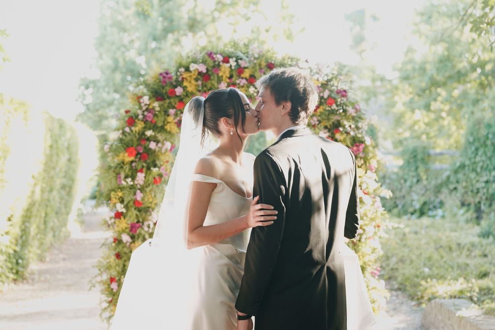 Bride in a white gown and groom in a dark suit share a kiss beneath a colorful floral arch at an outdoor garden wedding.