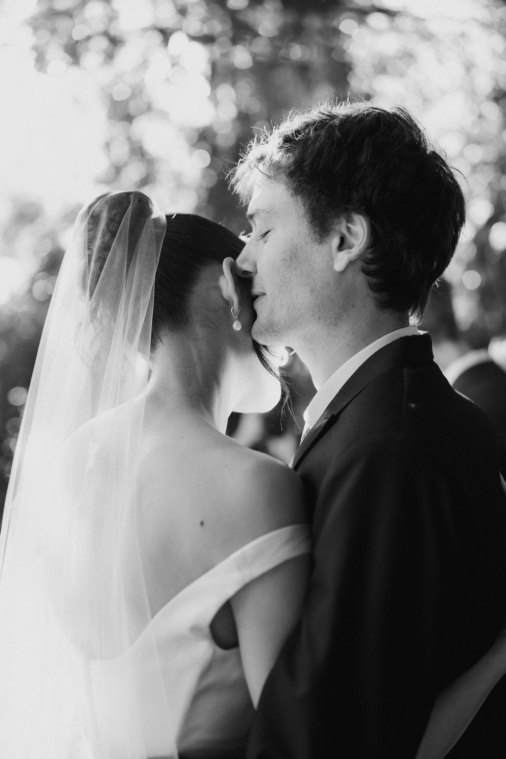 Black and white wedding portrait of a groom tenderly pressing his face to his veiled bride in an off-the-shoulder gown outdoors.