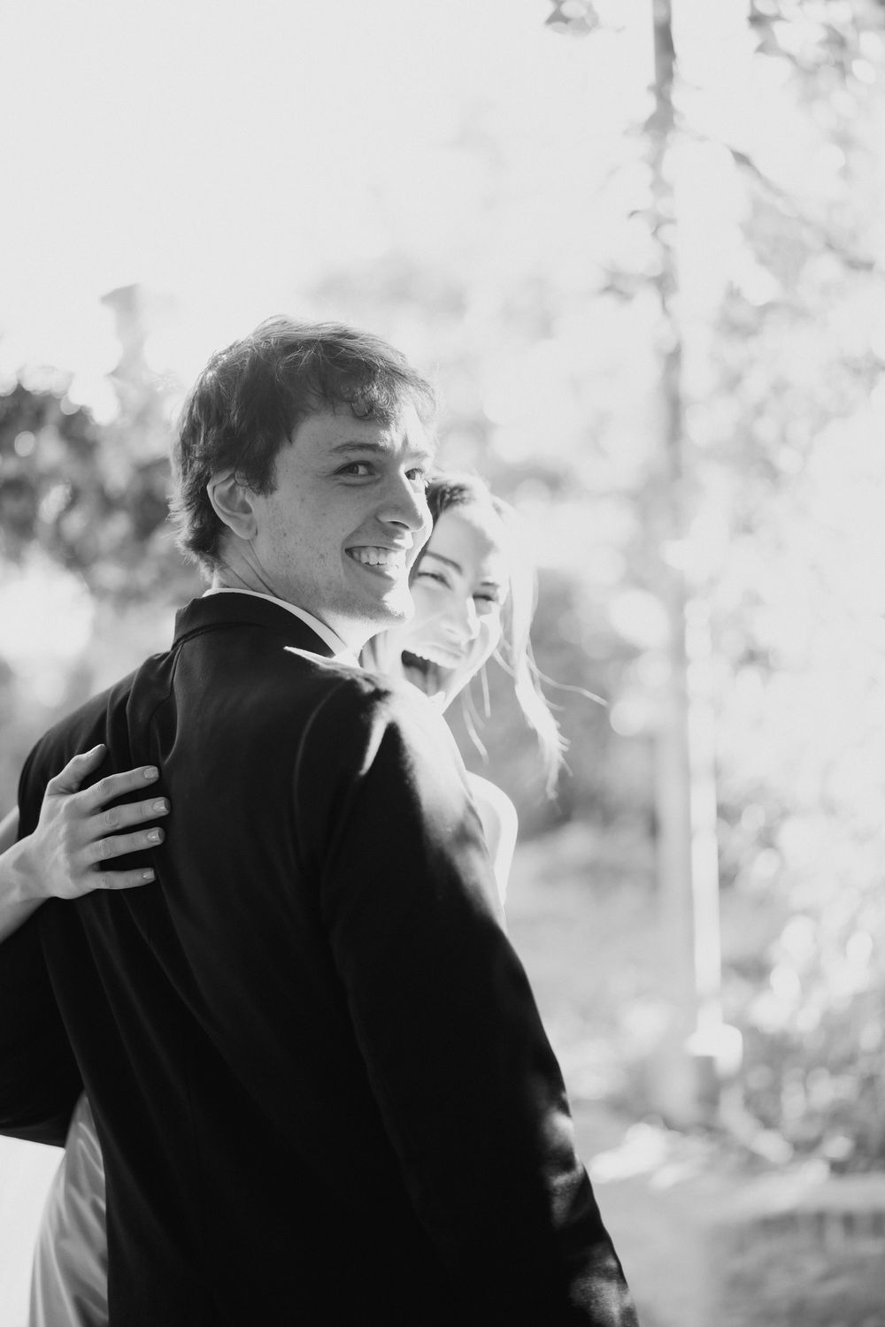 Black and white photo of a smiling man in a dark suit looking back as a laughing woman embraces him from behind outdoors.