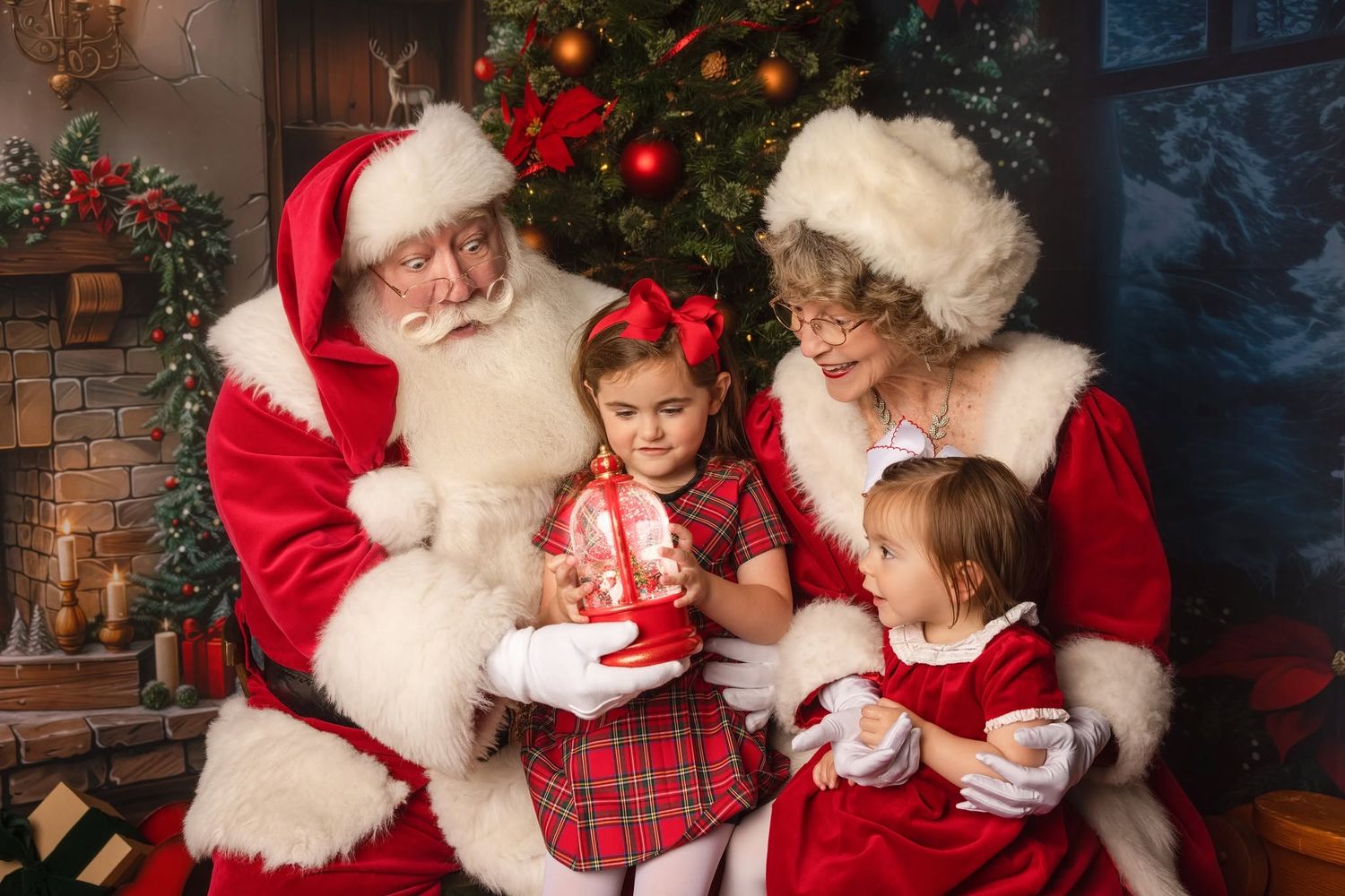Santa Claus and a woman in a fur-trimmed red coat pose with two little girls holding a glowing ornament beside a decorated Christmas tree.