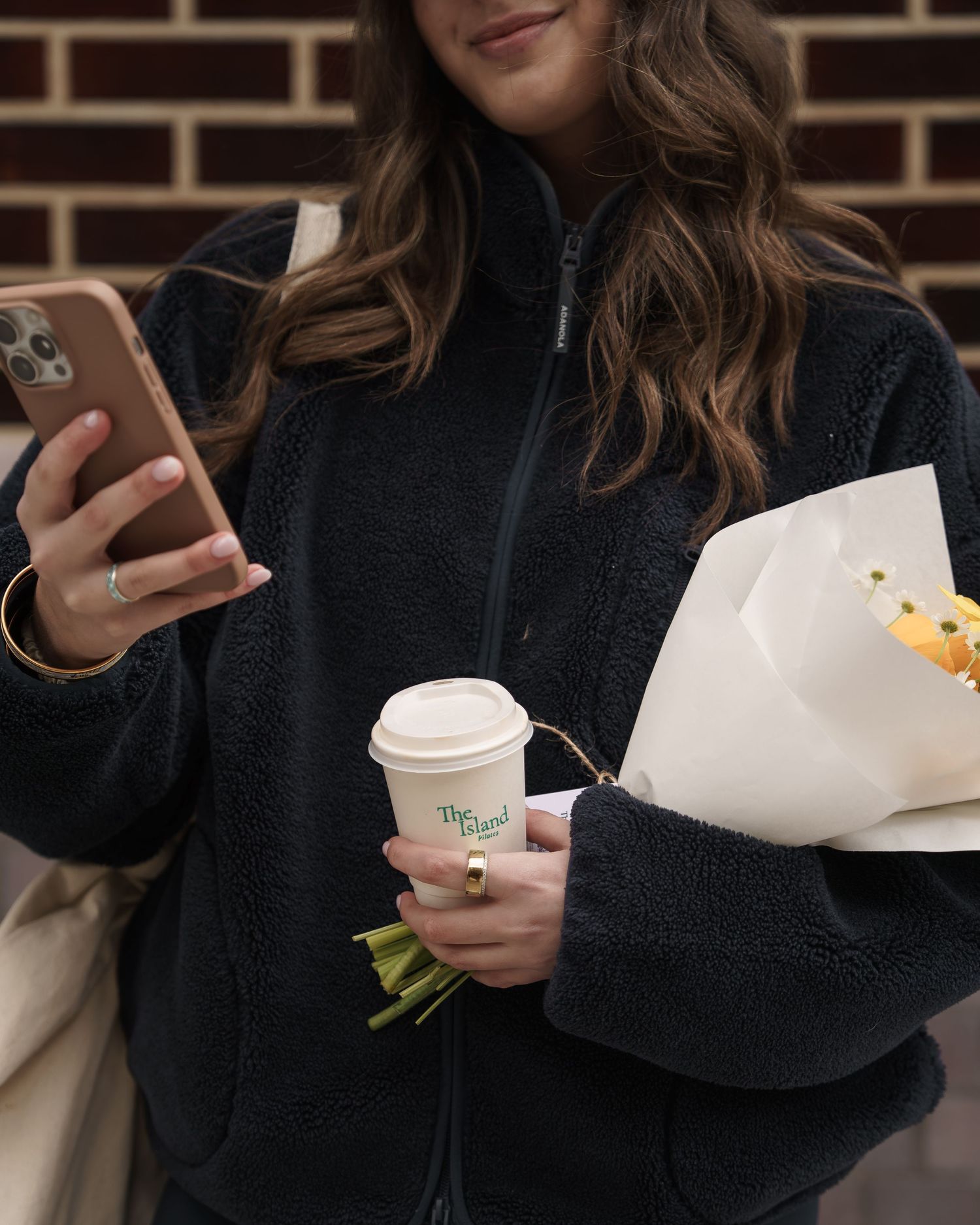 Woman in black fleece jacket holding coffee cup, flower bouquet, and phone while smiling outdoors.