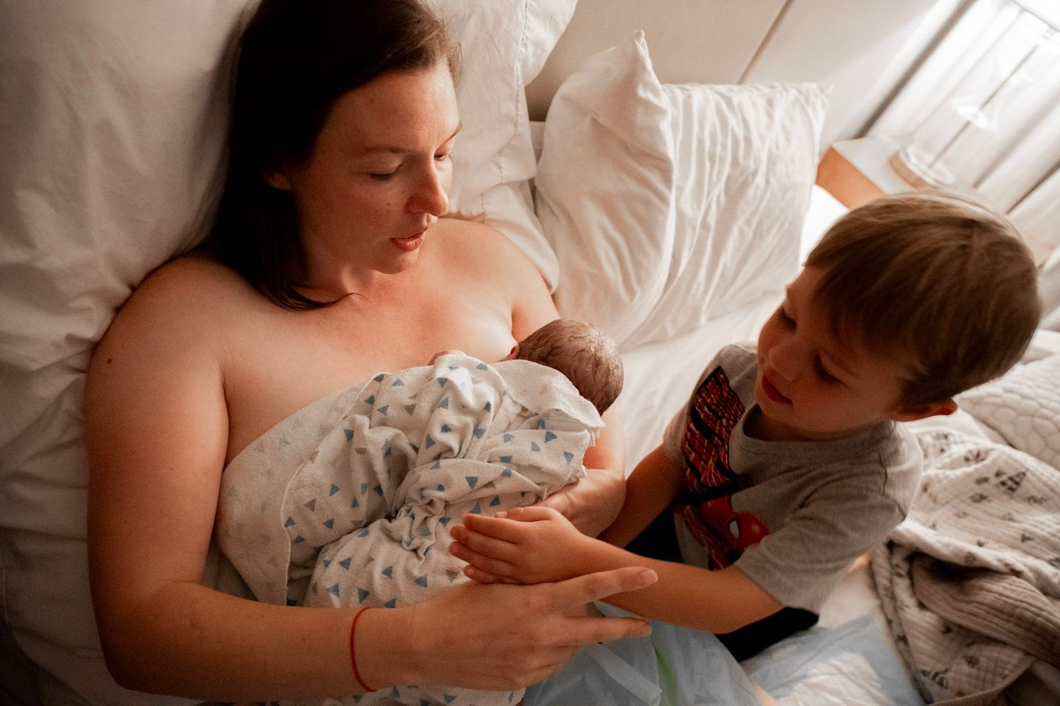 Mother resting in bed breastfeeding newborn baby while young toddler boy looks on with curiosity.