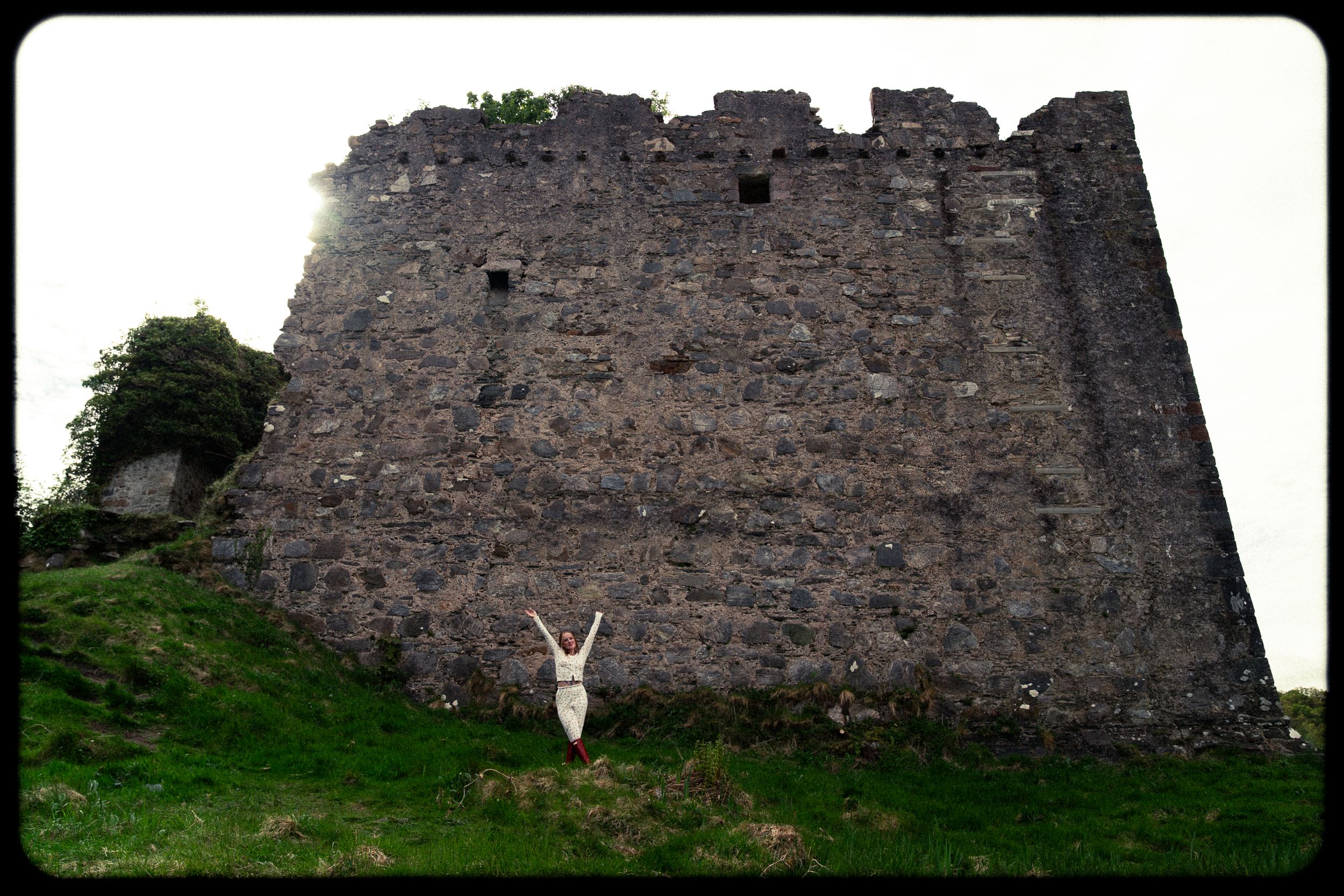 Woman with arms raised stands before ancient stone castle ruins on a grassy hill.