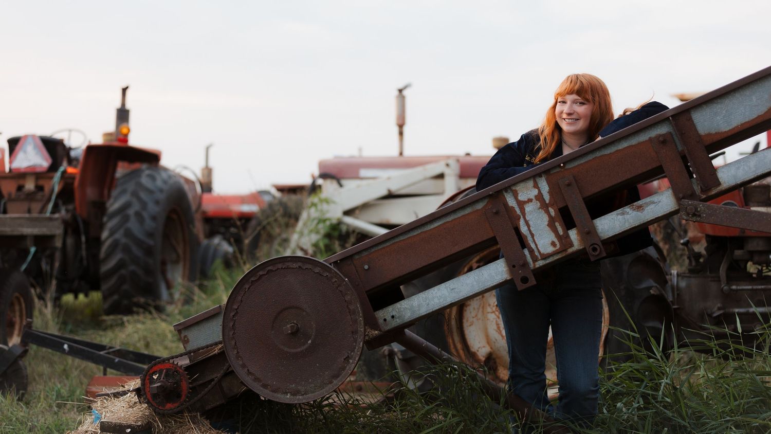Woman smiling among vintage red tractors and farm equipment in a rural junkyard setting.