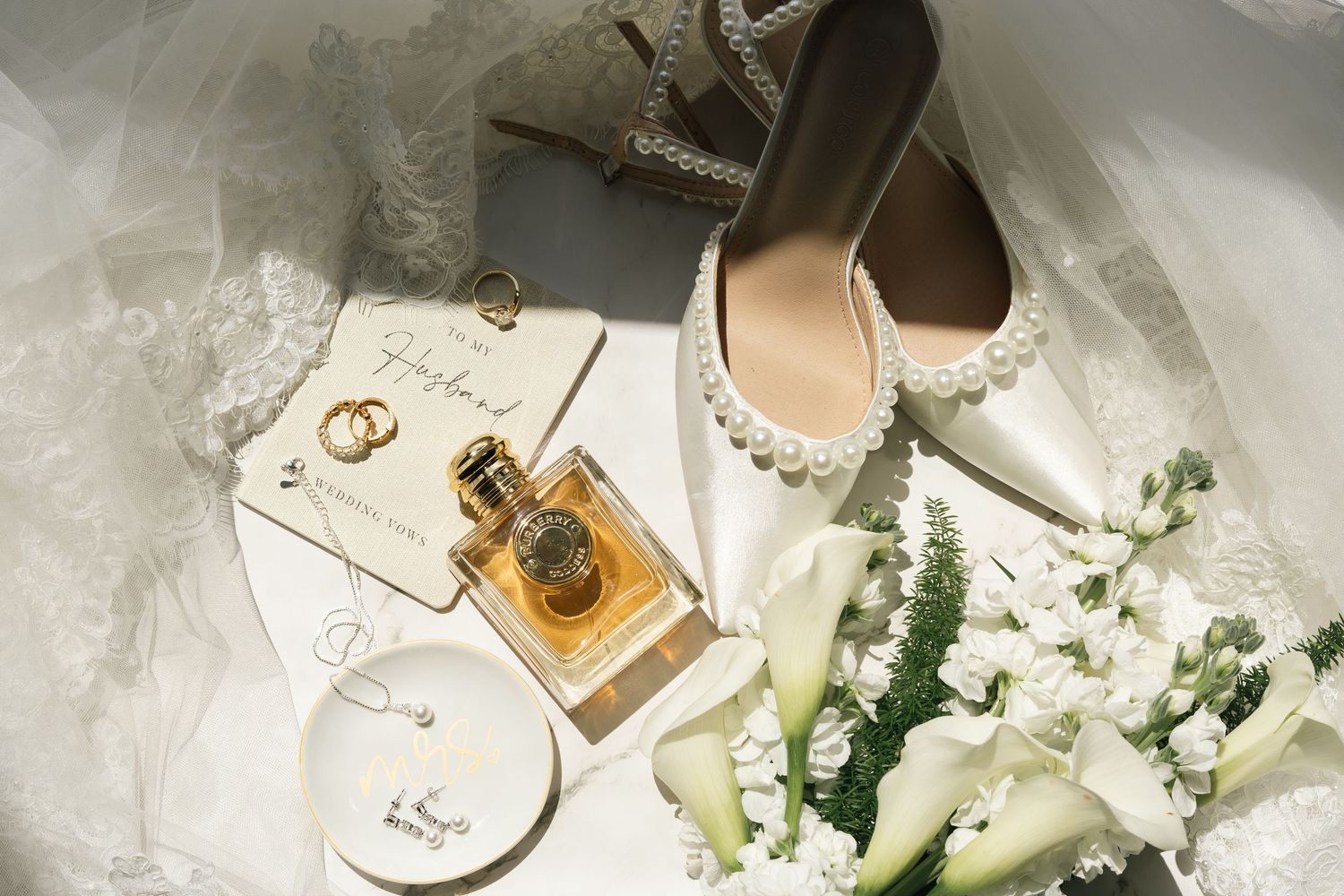 Elegant wedding flat lay featuring black oxford shoes, gold rings, YSL perfume, a watch, pearl heels, and white florals.