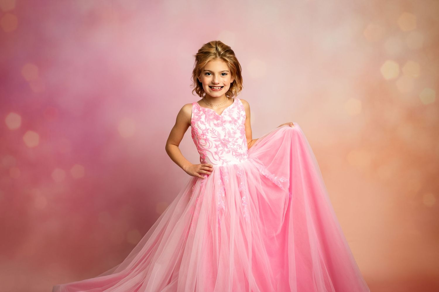 Young girl wearing a flowing pink princess ball gown with tulle skirt against a textured brown backdrop.