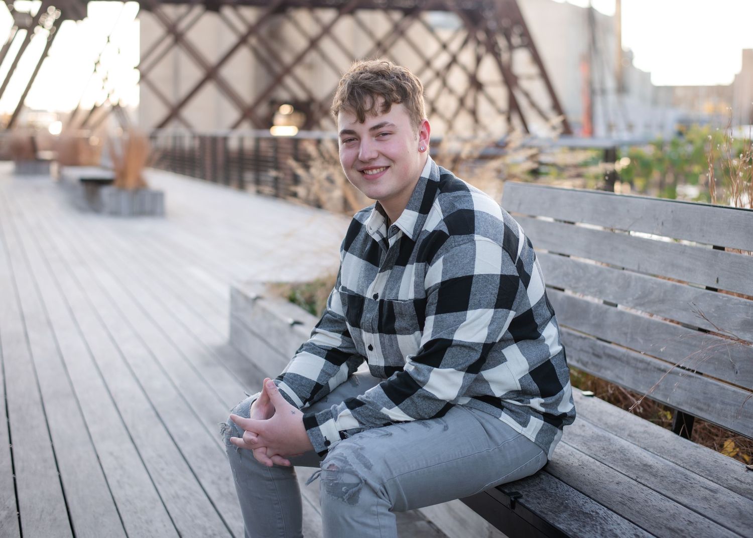 Smiling teenage boy in plaid shirt sitting on wooden bench outdoors near urban bridge structure.