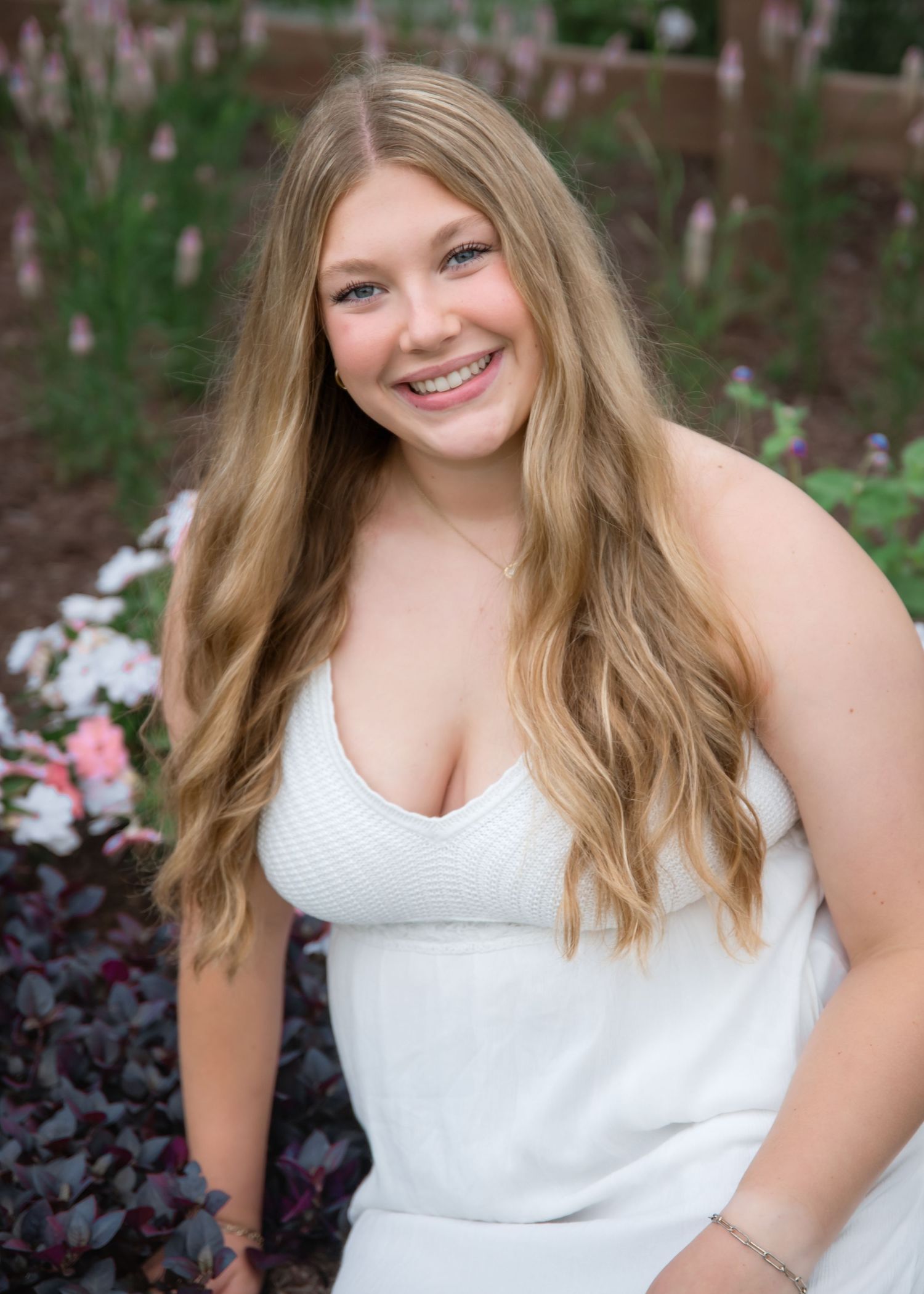 Smiling young woman with long blonde hair wearing a white dress, posing outdoors among colorful garden flowers.