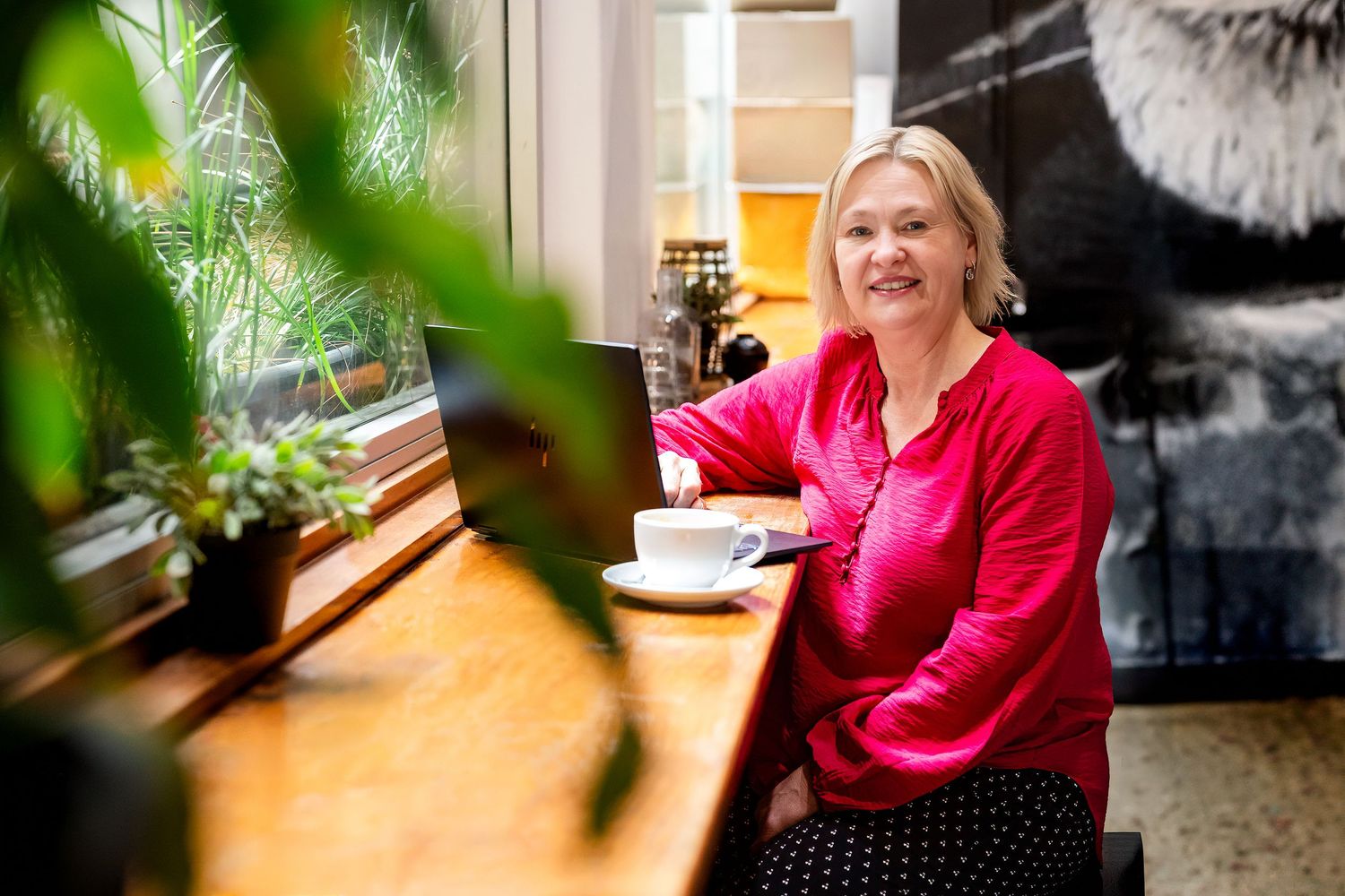 Woman in red blouse smiling at cafe table with coffee cup near window surrounded by green plants.