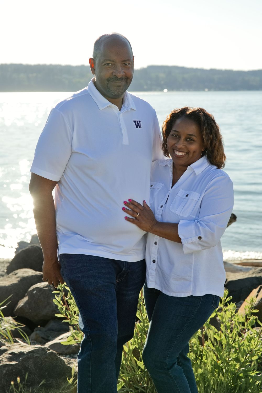 A smiling couple poses together near a rocky shoreline with a sunlit lake in the background on a sunny day.