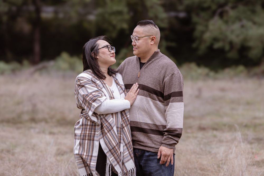 A couple shares a tender moment outdoors, smiling at each other in a grassy field surrounded by trees.