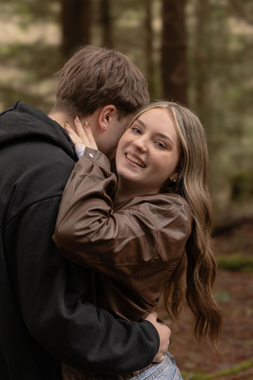 A smiling woman in a brown leather jacket embraces a man in a forest setting during an outdoor couples photo session.