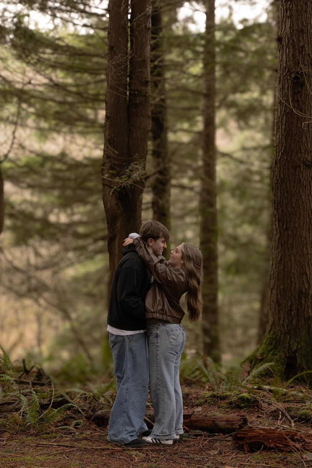 A young couple shares a romantic moment in a misty forest, surrounded by tall pine trees and mossy ground.