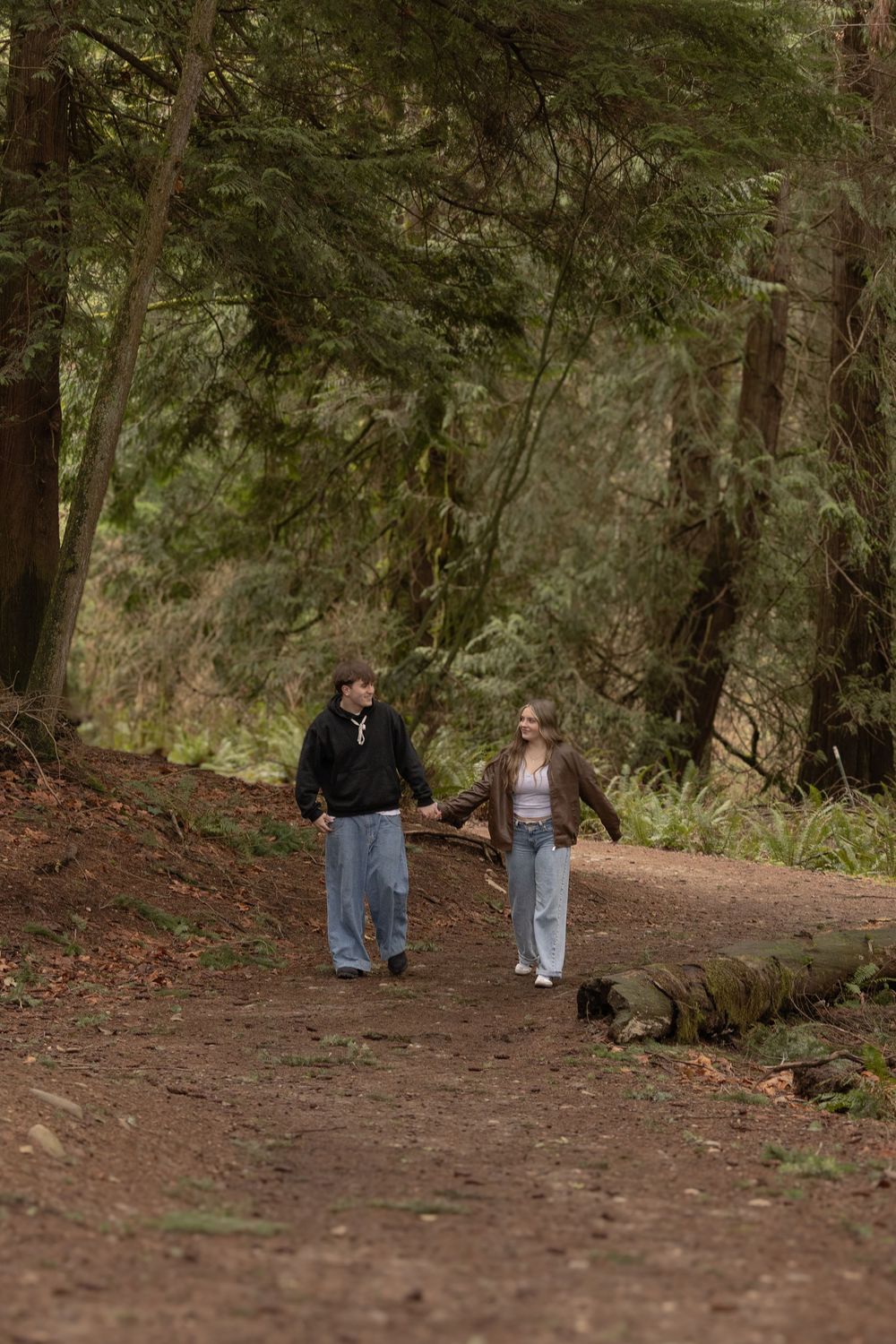 Two people walking together on a forest trail surrounded by tall trees and fallen logs.