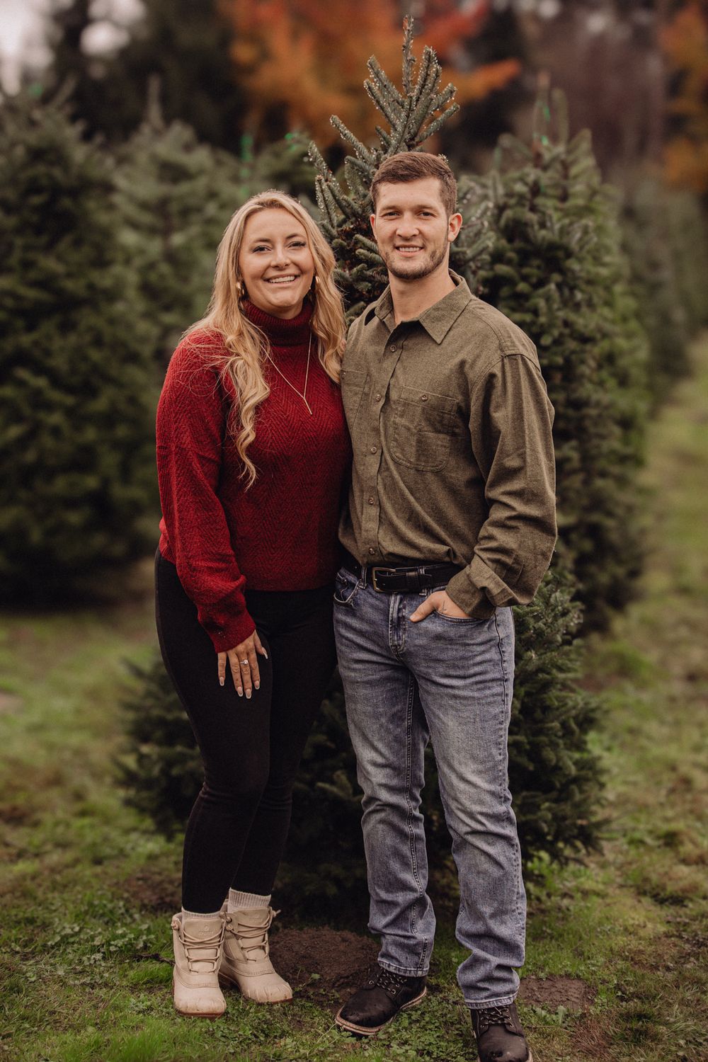Couple smiling together at a Christmas tree farm, woman in red sweater and man in olive shirt, surrounded by evergreen trees.