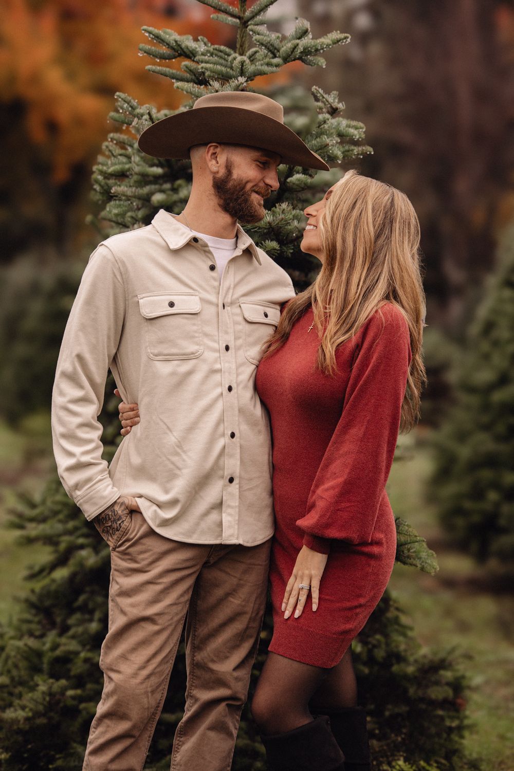 Couple embracing among Christmas trees, man in tan cowboy hat and shirt, woman in red dress, autumn outdoor setting.