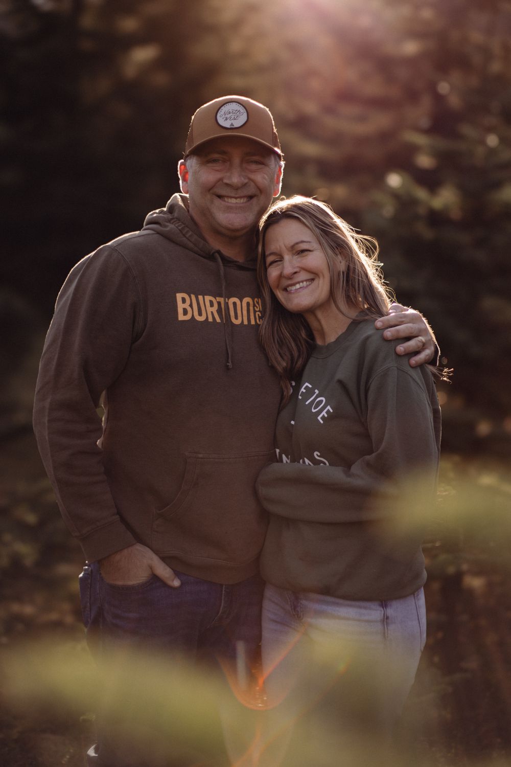 Couple smiling outdoors in golden sunlight, man in Burton hoodie and cap, woman in green sweatshirt, embracing in nature.