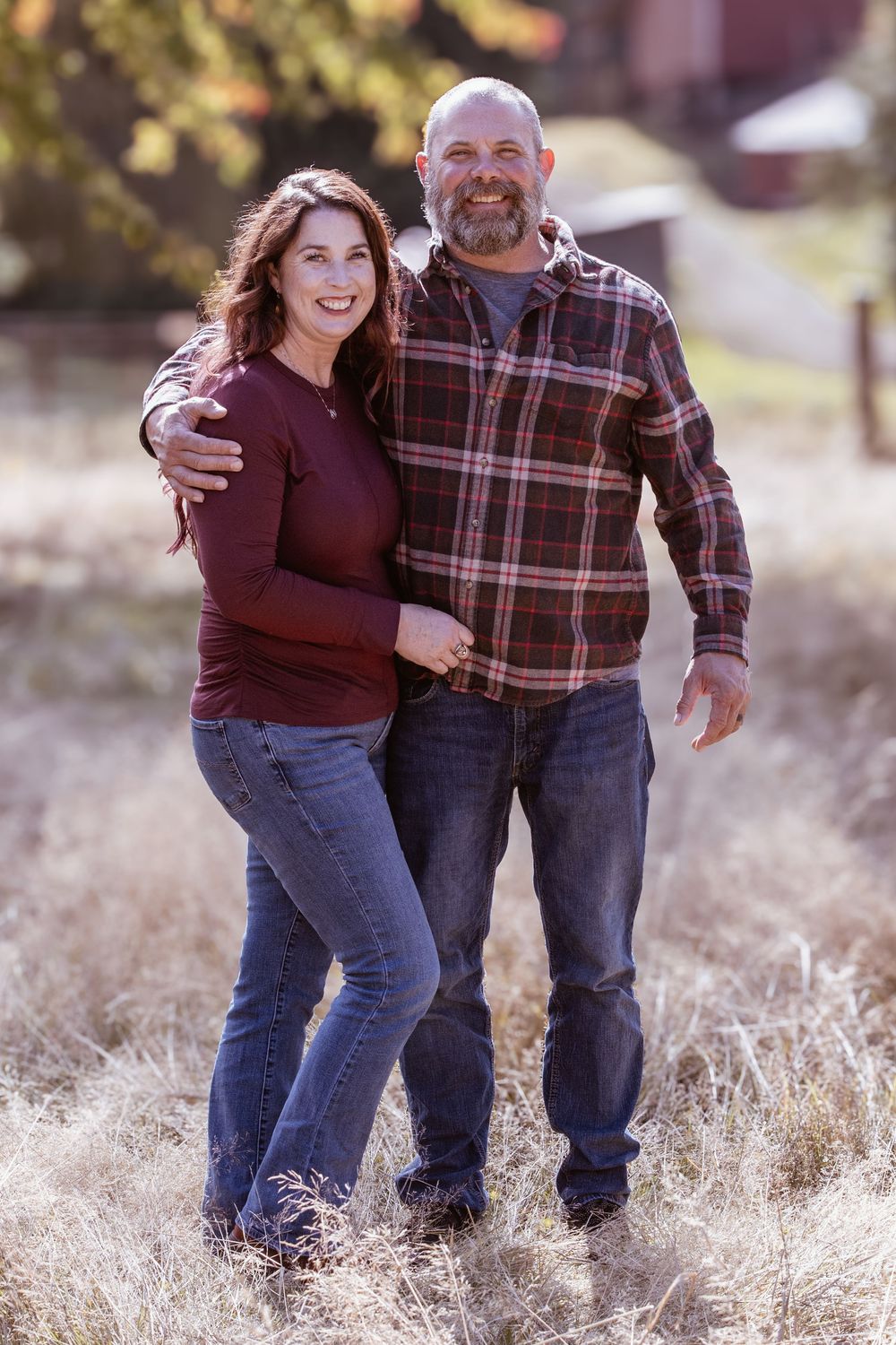 A smiling couple poses outdoors in a field, wearing coordinating burgundy and plaid flannel shirts with jeans on a sunny fall day.