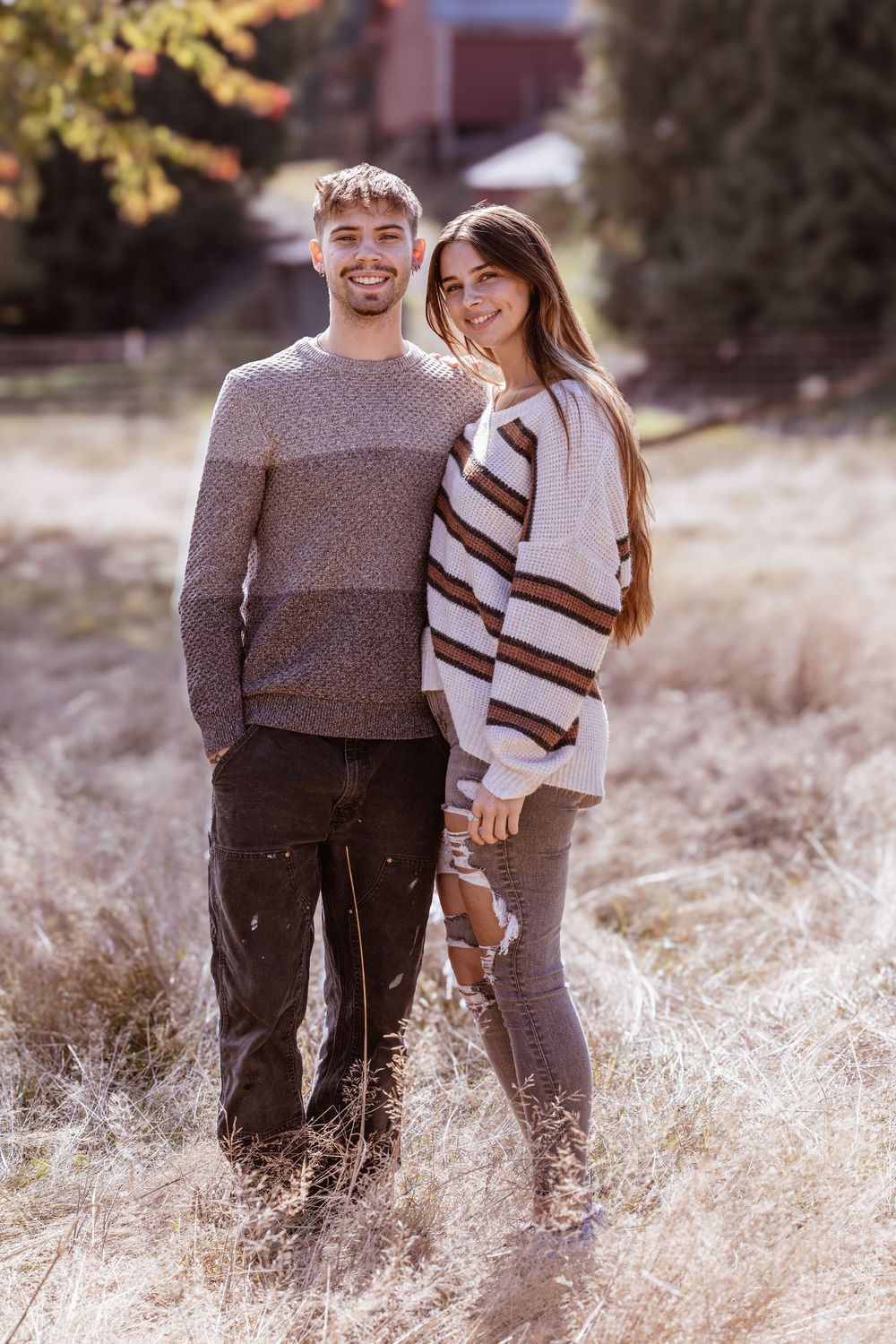 A smiling couple poses together in a golden field wearing cozy sweaters on a sunny autumn day.