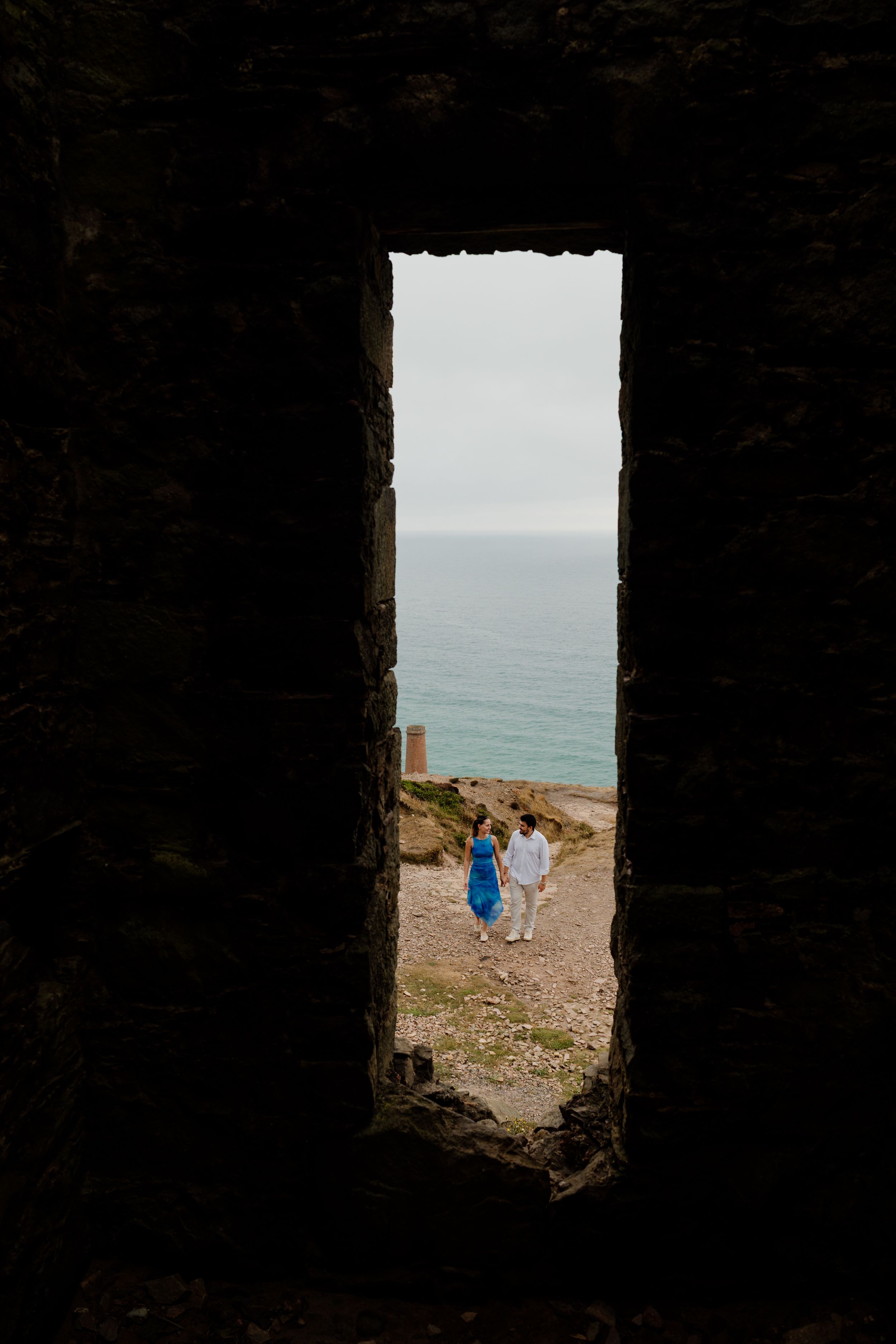 Couple walking toward the sea viewed through a narrow stone archway opening on a rocky coastal path.