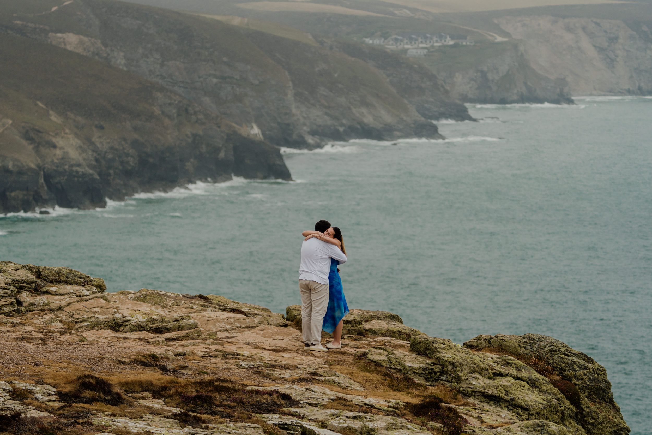 Couple embracing on rocky coastal cliffs overlooking turquoise ocean waters with dramatic green headlands in background.