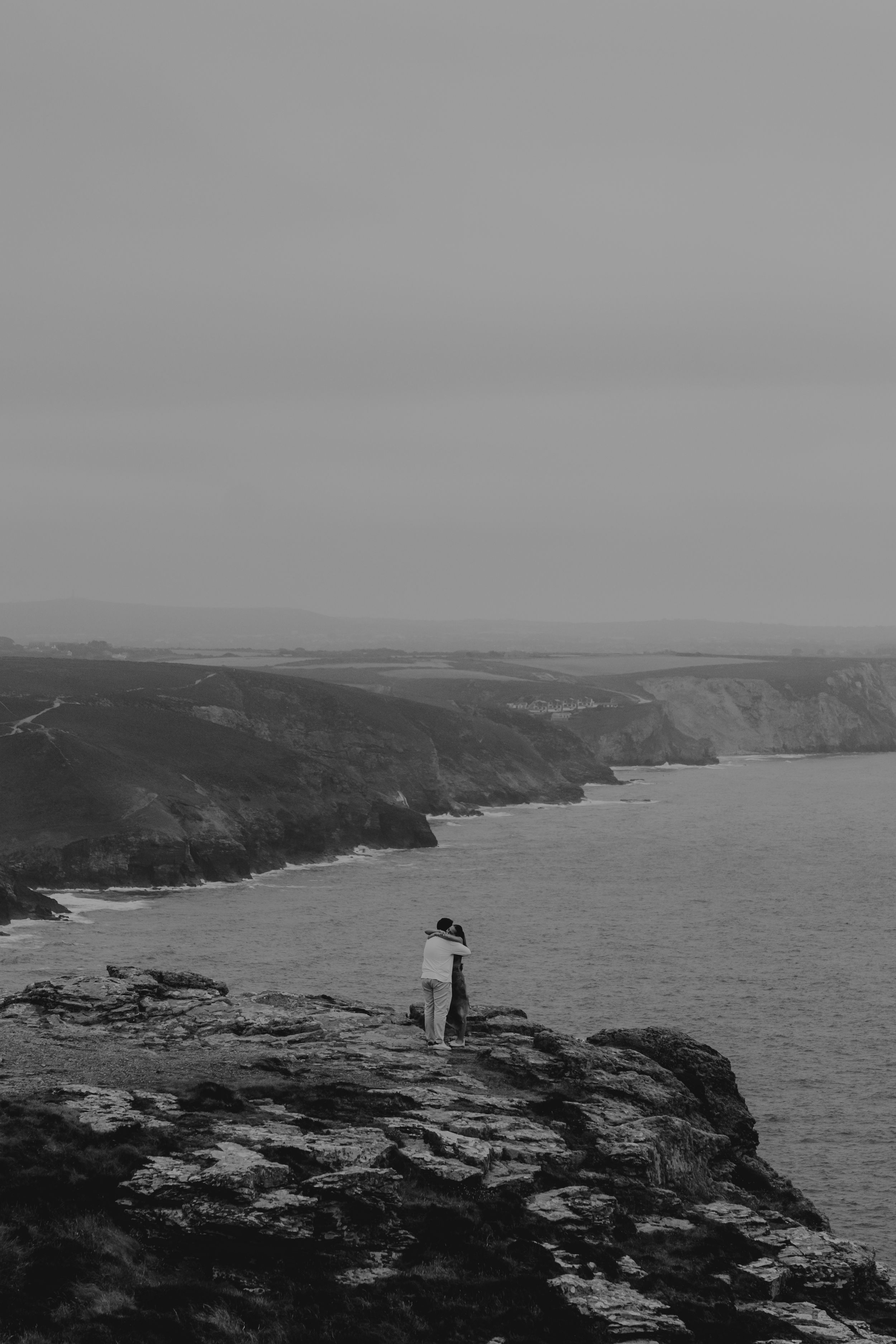 Black and white photo of a lone figure standing on coastal cliffs overlooking a dramatic rocky shoreline.