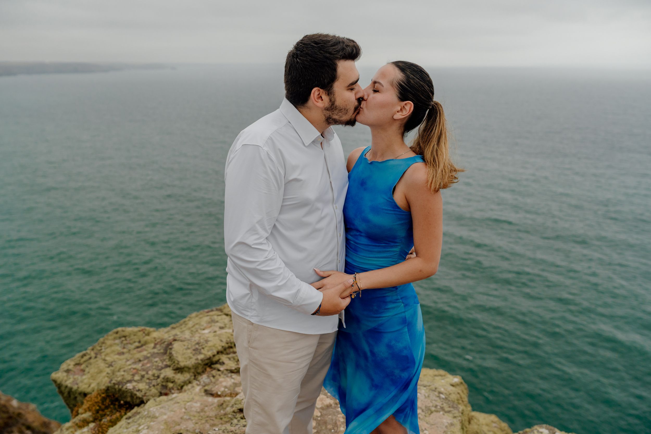Couple kissing on rocky coastal cliff with turquoise ocean backdrop, woman in blue dress, man in white shirt.