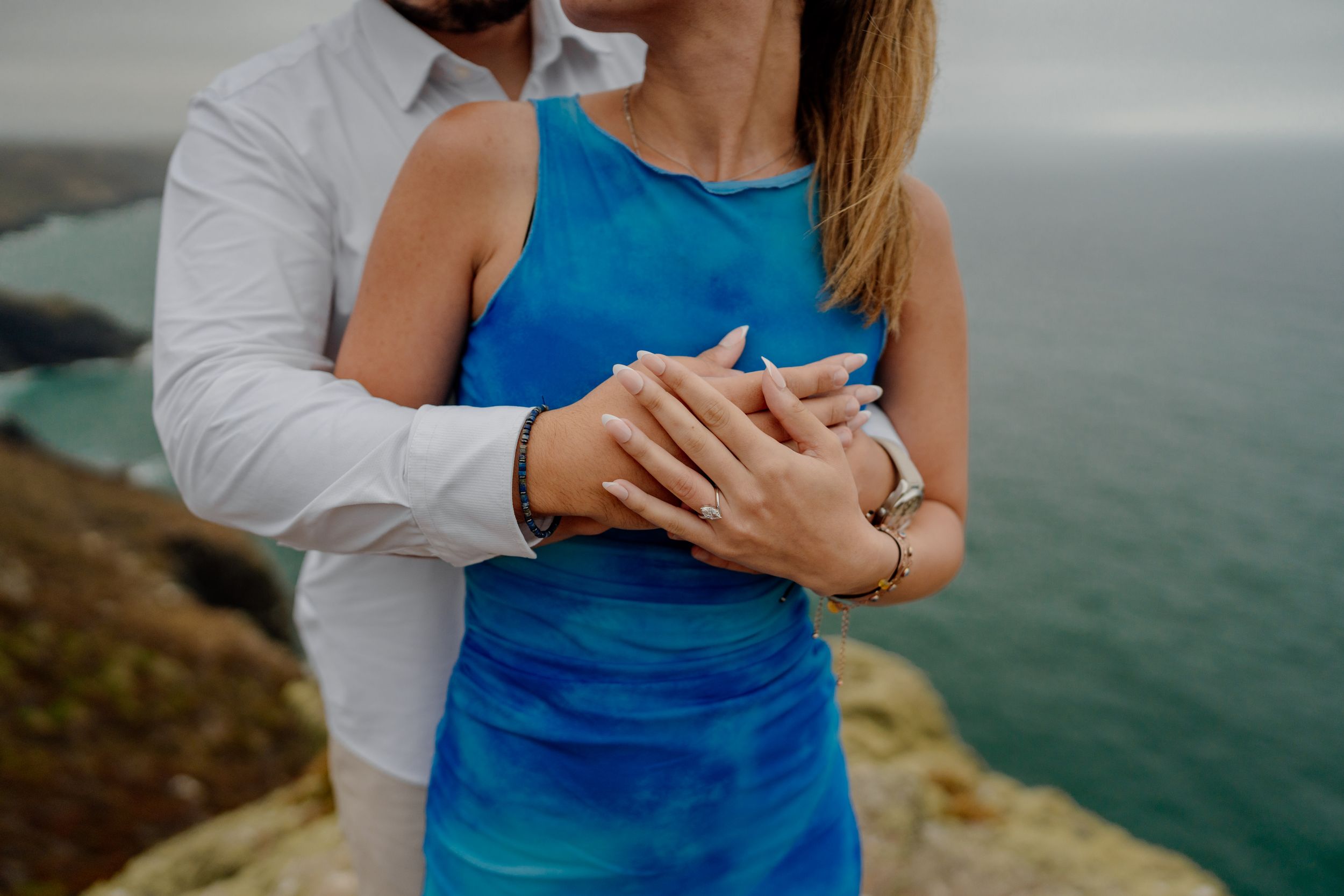 Couple embracing on coastal cliffs, woman in blue dress showing engagement ring, ocean backdrop.