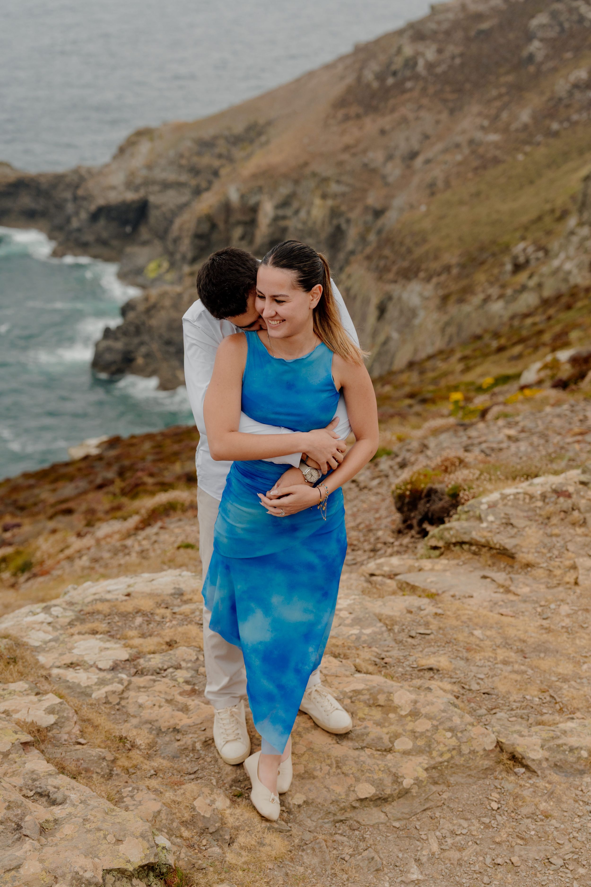 Couple embracing on rocky coastal cliffs, woman in blue dress smiling, ocean waves visible in background.