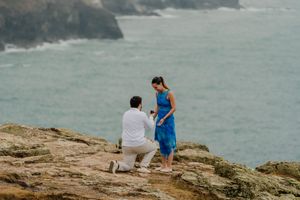 Man proposes on rocky coastal cliff to woman in blue dress with ocean backdrop.