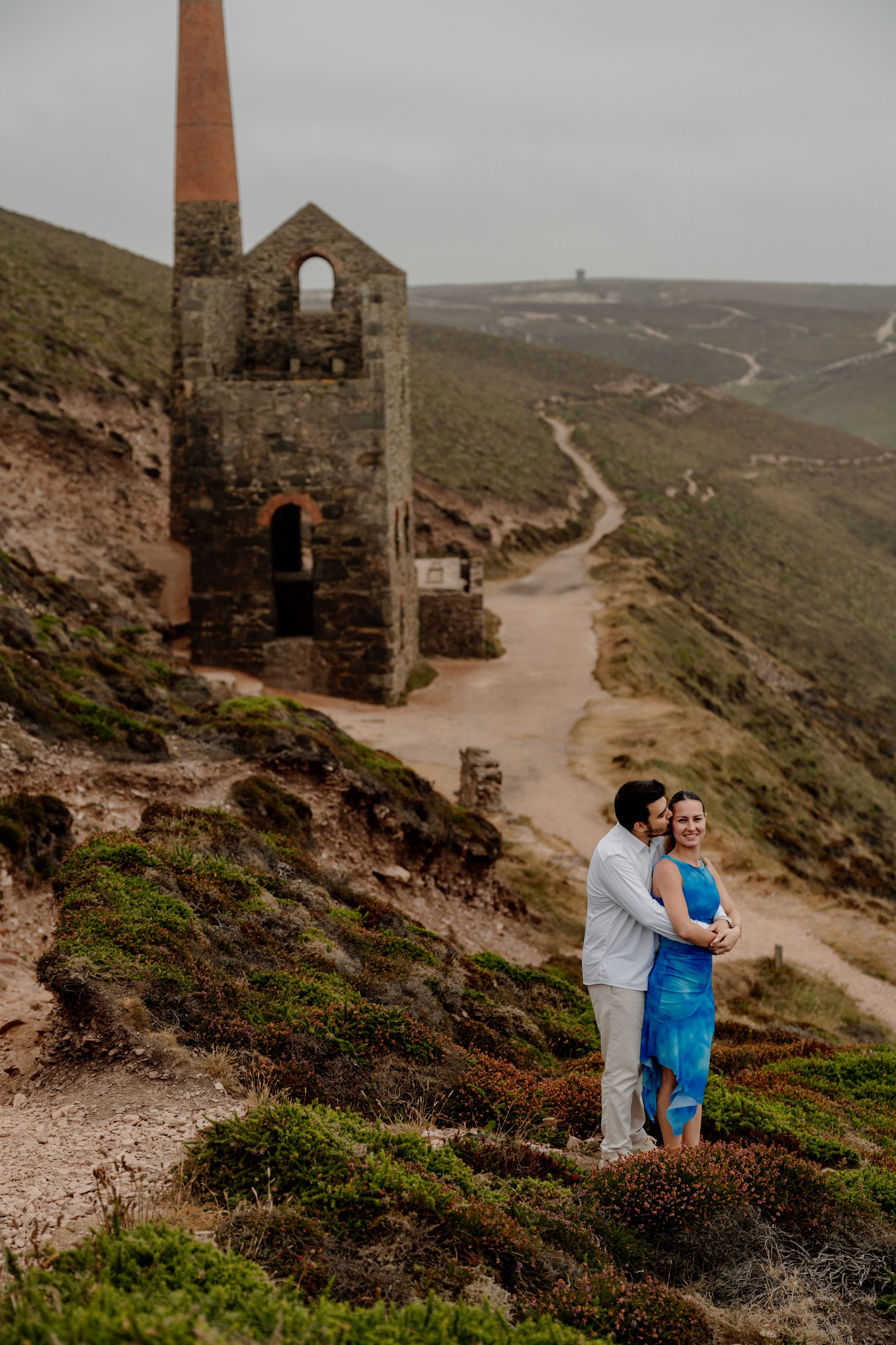 Couple embracing near historic stone engine house ruins on a coastal hillside in Cornwall.