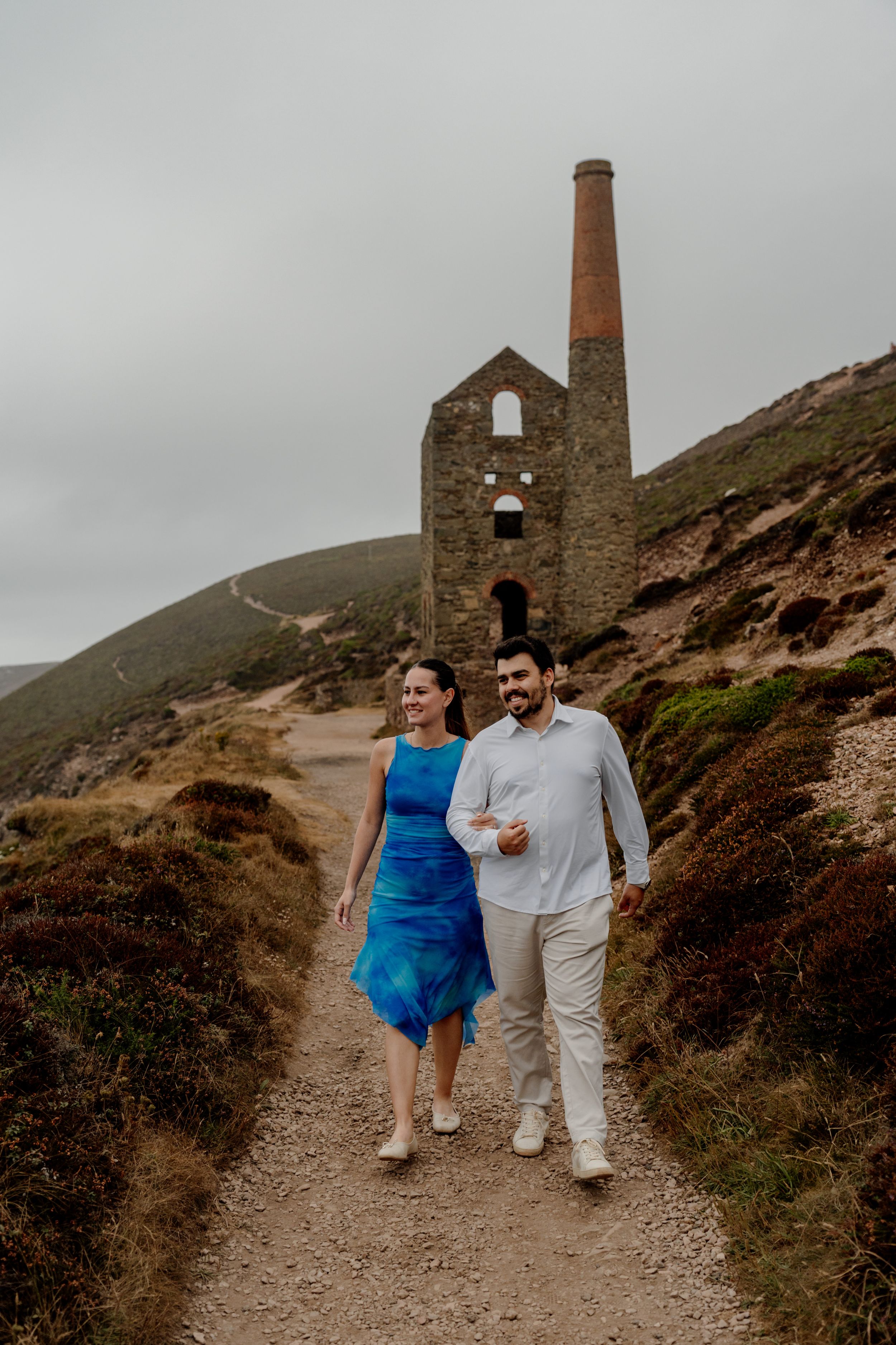 Couple walking hand in hand on a coastal path near a historic Cornish tin mine engine house on a cloudy day.