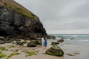 Couple embracing on a rocky beach with mossy boulders, ocean waves, and dramatic cliffs under an overcast sky.