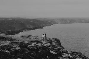 Two people stand on rocky coastal cliffs overlooking a dramatic shoreline in a black and white landscape photo.
