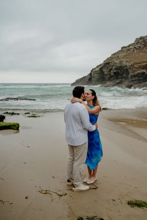Couple sharing a romantic kiss on a sandy beach with ocean waves and rocky cliffs in the background.