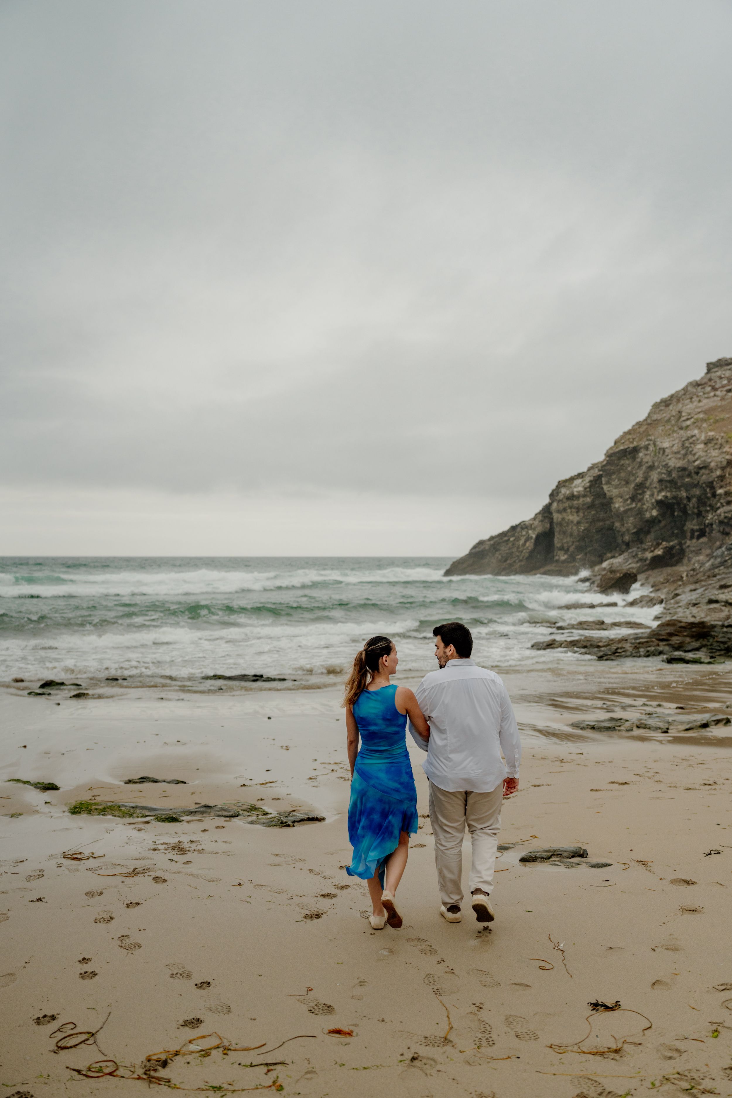 Couple walking hand in hand on a sandy beach with rocky cliffs and ocean waves under an overcast sky.