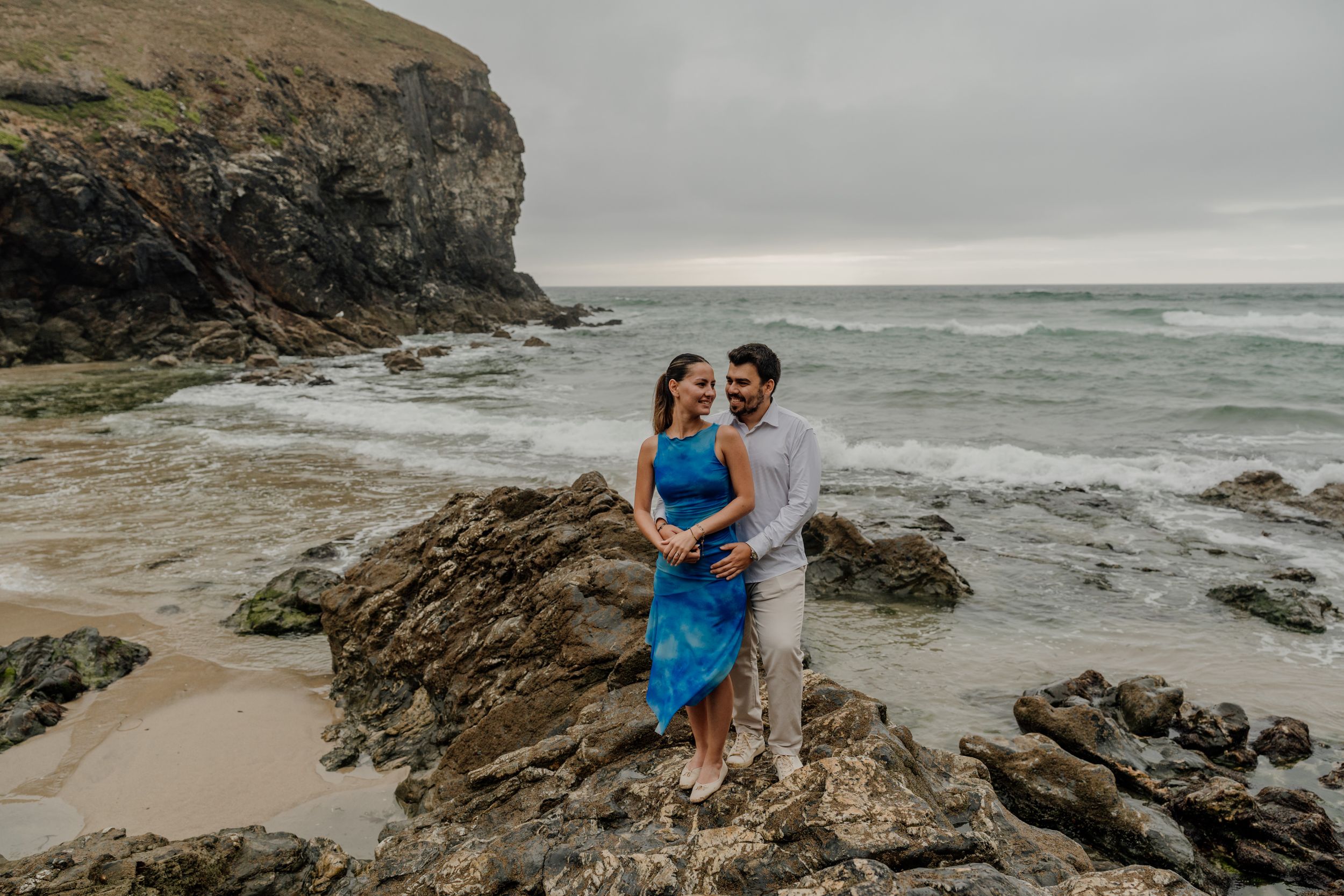Couple embracing on rocky beach with ocean waves and dramatic cliffs in background, woman in blue dress.