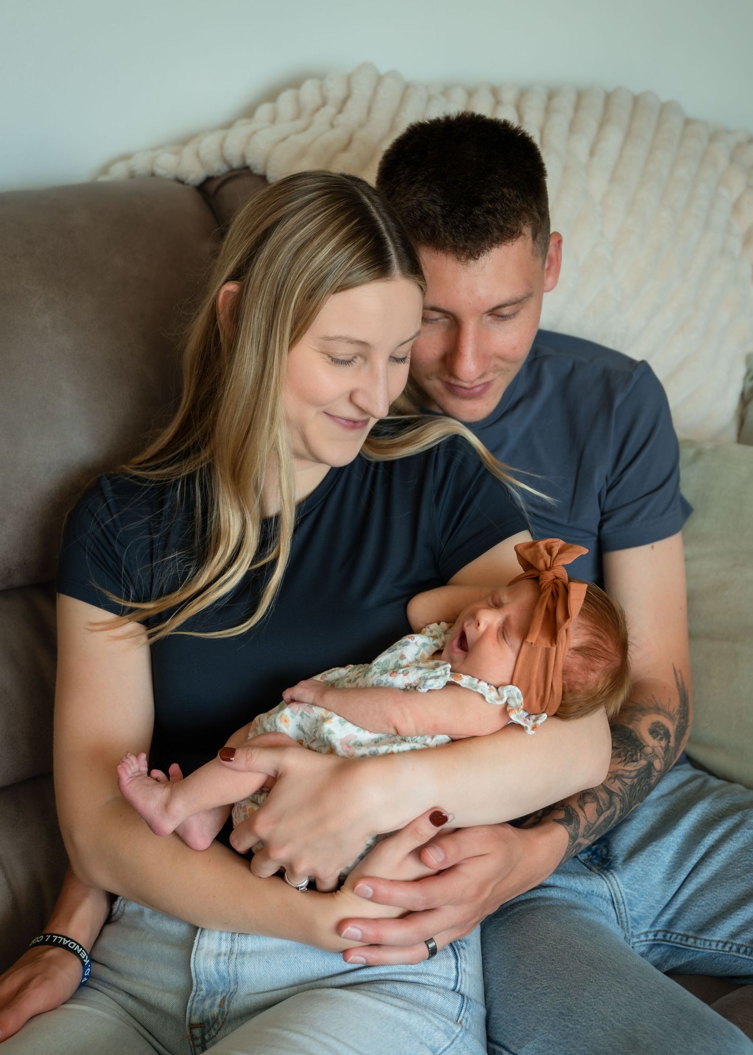 A young couple sits on a couch, lovingly gazing at their newborn baby girl wearing a floral outfit and bow headband.