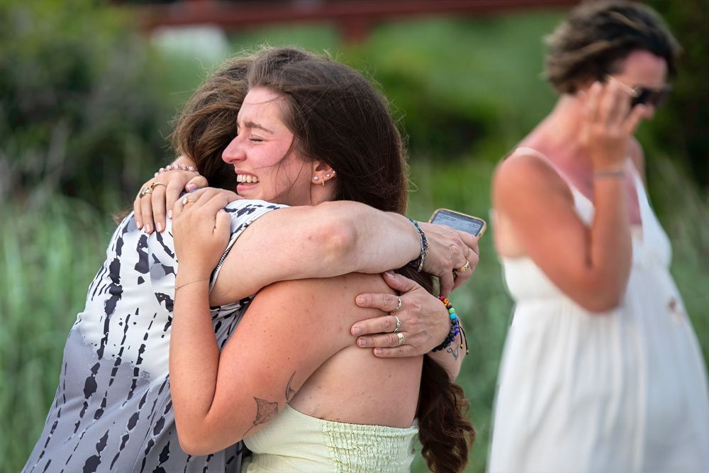 Two young women share a warm embrace outdoors while another woman stands nearby, capturing a joyful reunion moment.