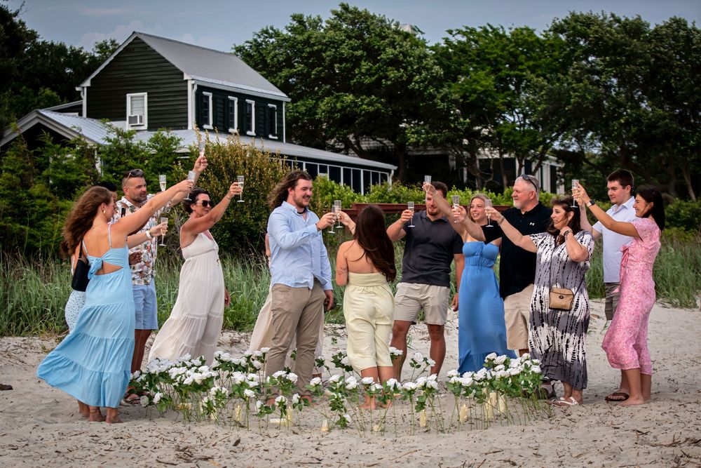 Group of friends toasting on a beach near a house, celebrating outdoors with flowers on the sand.