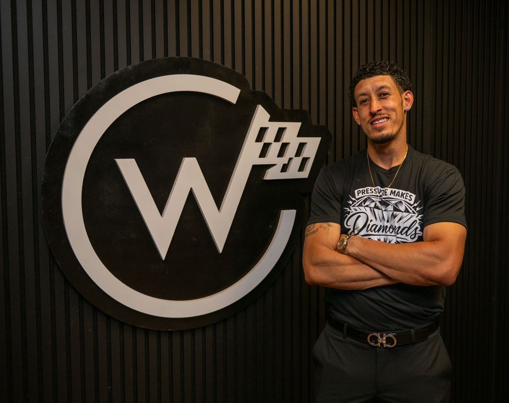 Man in graphic tee posing confidently beside a large W logo sign on a dark paneled wall.